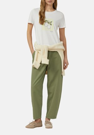 Femme portant un t-shirt blanc avec un motif citron, un pull beige en maille noué autour de la taille, un pantalon vert olive et des mocassins beiges.