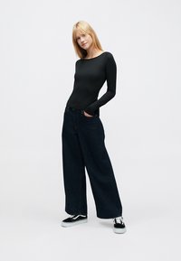 Črno bodi z dolgimi rokavi, kombinirano z visokim pasom širokih temnih jeans hlač in črnimi supergami z belim podplatom. Enobarvno ozadje.