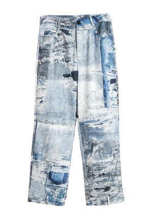 Joven de pie, vestido con chaqueta y pantalones de patrón abstracto azul y blanco, con zapatos color beige, sobre un fondo blanco.