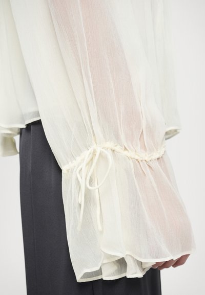 Kerge, poolläbipaistev valge pluus kogutud varrukatega, mis on käeõrtes nööriga kokku tõmmatud, pehme ja voolava kangaga.