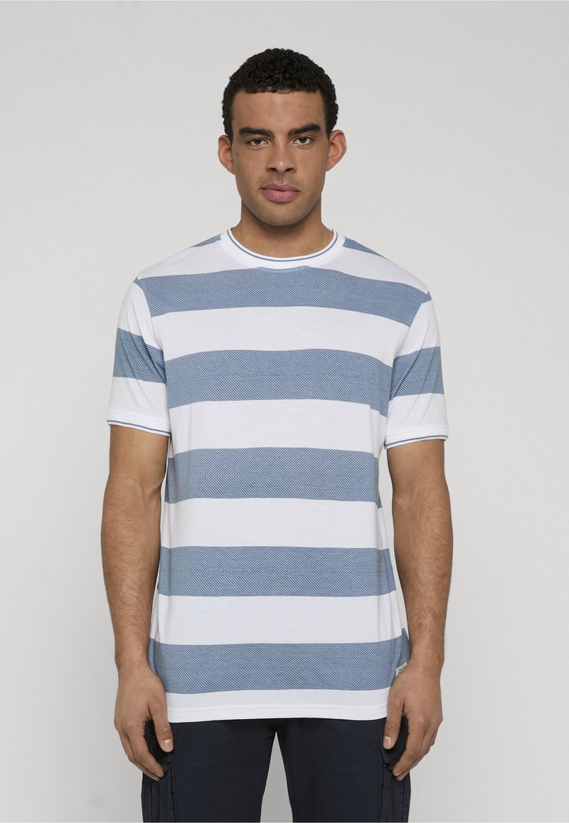 Man die naar voren kijkt, draagt een wit en blauw horizontaal gestreept T-shirt met korte mouwen en donkere broek tegen een egale achtergrond.