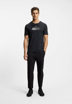 Junger Mann steht, trägt ein schwarzes T-Shirt mit Grafik und kurzen Ärmeln, schwarze schmale Hosen und schwarze Sneakers vor einfachem hellem Hintergrund.