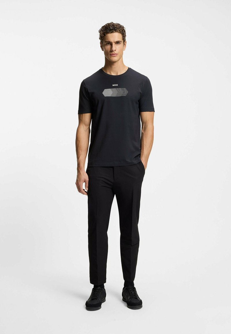 Jonge man staand, draagt een zwarte T-shirt met korte mouwen en grafisch ontwerp, zwarte slim-fit broek en zwarte sneakers tegen een effen lichte achtergrond.