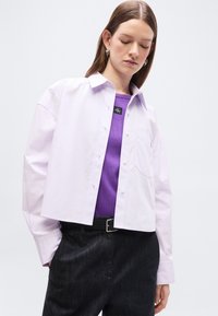 Γυναίκα με κοντό λευκό πουκάμισο με κουμπιά πάνω από μωβ τοπ, μαύρη ζώνη και σκουρόχρωμο παντελόνι ψηλής μέσης, που κοιτάει προς τα κάτω.