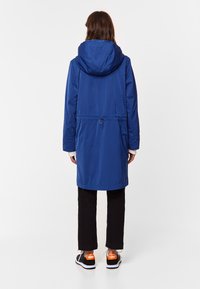 Blå vattentät parka med dragsko i midjan, huva och långa ärmar, kombinerad med svarta byxor och sneakers med oranga detaljer.