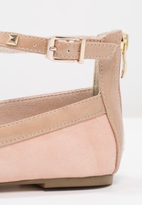 Chaussure plate en daim beige avec bride à la cheville, boucle dorée et détail clouté, avec fermeture éclair à l’arrière.