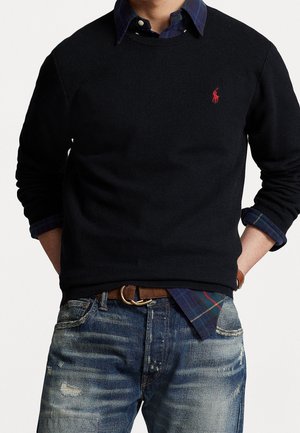 Mand iført sort striktrøje med rødt logo over blå ternet skjorte og slidte blå jeans med brun bælte, hænderne i lommerne.