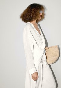 Witte linnen blouse met een gedraaide voorkant, gecombineerd met bijpassende losse broek. Houdt een gestructureerde beige geweven tas vast en draagt een zonnebril.