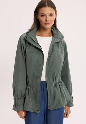 Kobieta ubrana w zieloną kurtkę na zamek z regulowanym talią i mankietami, nałożoną na białą koszulę i niebieskie spodnie, stojąca na jednolitym tle.