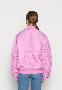 Kvinna med brunt hår i hästsvans, bärande stora örhängen, en ljusrosa rynkad bomberjacka och blå jeans, visad bakifrån.