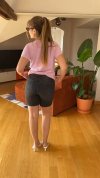 Vaaleanpunainen istuva lyhythihainen paita, mustat farkkushortsit, joissa on kaksi takataskua, ja valkoiset avokkaat. Sisätila, jossa on kasvi.