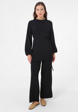 Kvinde iført en sort tunika med lange ærmer og matchende vide bukser samt beige loafers, accessorized med guldbelagte øreringe og en perlekæde.