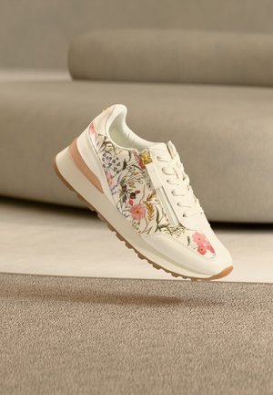 Baskets blanches avec motif floral rose, fermeture éclair latérale, lacets blancs et semelle marron clair, posées sur un tapis beige près d'un canapé beige.