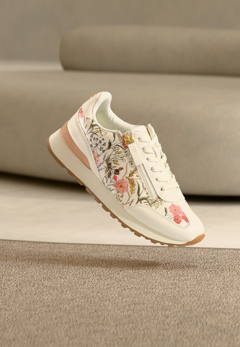 Baskets blanches avec motif floral rose, fermeture éclair latérale, lacets blancs et semelle marron clair, posées sur un tapis beige près d'un canapé beige.