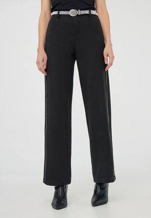 Donna che indossa pantaloni larghi neri con strisce bianche laterali, una cintura bianca e stivali neri alla caviglia su uno sfondo neutro.