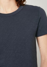 Tums zilā īsām piedurknēm krekls, izgatavots no mīksta, teksturēta auduma. Tam ir klasiskā apaļā dekoltē un minimāla šuvju detaļa.