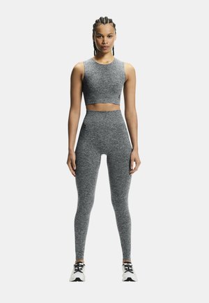 Donna in piedi, rivolta verso l'obiettivo, che indossa un top corto grigio senza maniche, leggings a vita alta abbinati e scarpe da ginnastica bianche, su sfondo bianco.