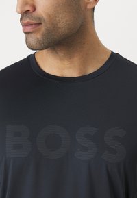Fekete, rövid ujjú póló kerek nyakkal, amelyen diszkrét vízszintes csíkozásban a "BOSS" szó hangsúlyosan szerepel az elején.
