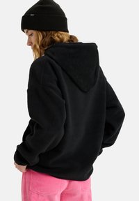 Felpa nera in pile con un grande cappuccio, vestibilità comoda e superficie testurizzata, abbinata a pantaloni di velluto a coste rosa. Berretto nero sulla testa.