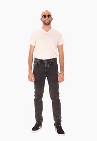 Homme avec tête chauve, barbe et lunettes de soleil, portant un t-shirt blanc à col en V, un jean noir et des baskets noires, se tenant contre un fond blanc.