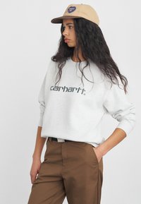 Jasnoszara bluza z napisem "carhartt", okrągły dekolt, ściągacze na rękawach. Brązowe spodnie z bocznymi kieszeniami, noszone z beżową czapką z logo.