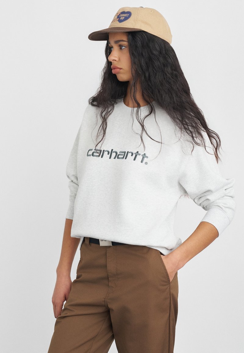 Jasnoszara bluza z napisem "carhartt", okrągły dekolt, ściągacze na rękawach. Brązowe spodnie z bocznymi kieszeniami, noszone z beżową czapką z logo.