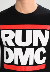 Černé bavlněné tričko s výrazným bílým textem "RUN" nad "DMC" v červeném obdélníku. Klasický design s kulatým výstřihem, hladká textura.