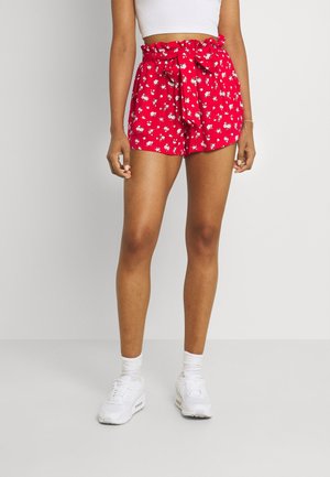 Vrouw draagt rode shorts met hoge taille en witte bloemenprint, een gestrikte riem, een wit cropped topje, witte sokken en witte sneakers, staand op een grijze vloer.