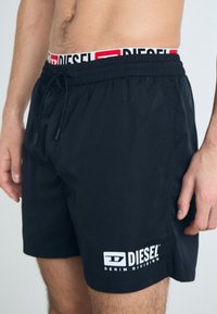 Black swim shorts with an elastic waistband featuring a white and red "DIESEL" logo. Made from lightweight fabric, with an adjustable drawstring.