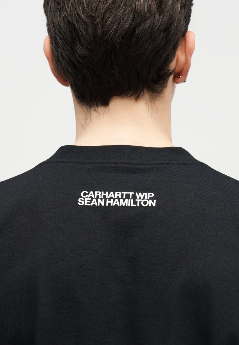 Person von hinten gesehen, trägt ein schwarzes Hemd mit der Aufschrift "CARHARTT WIP SEAN HAMILTON" in Weiß auf dem oberen Rücken.