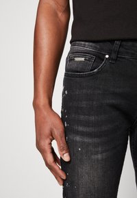 Mustat denimit housut, kapealla leikkauksella, joissa on kevyttä kulutusta ja hopeinen merkki vyötäröllä. Sileä pinta, hienovaraiset haalistumisefektit.