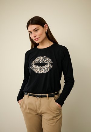 Schwarzes Langarmshirt mit einem Leopardenmotiv und Kussgrafik. Getragen mit khakifarbenen Hosen und einem schwarzen Gürtel. Glattes Material, figurbetonender Schnitt.