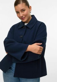 Ženska v mornarsko modrem plašču z gumbi in širokimi rokavi prekrsti roke, nežno se smehlja na enostavni belem ozadju.