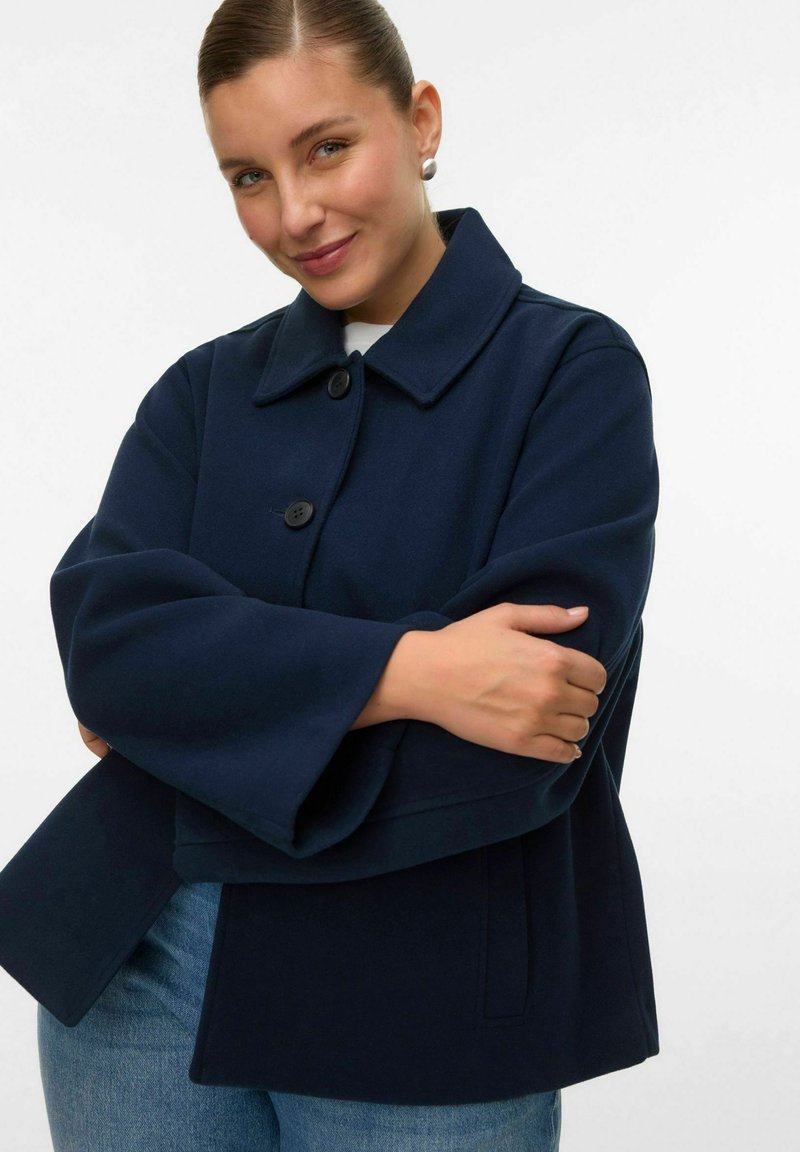 Ženska v mornarsko modrem plašču z gumbi in širokimi rokavi prekrsti roke, nežno se smehlja na enostavni belem ozadju.