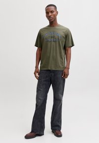 Olivengrøn bomulds T-shirt med blå grafisk tekst, kombineret med mørkegrå vidde jeans. Brune sko og enkle ørestikker bæres.