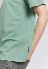 Camiseta de algodón verde claro con mangas cortas, corte recto y una etiqueta lateral con el texto de la marca. La textura parece suave y casual.