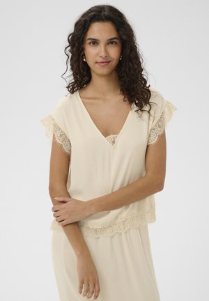 Mladá žena s kučeravými tmavými vlasmi, oblečená v krémový top a sukne s čipkou, stojaca so založenými rukami pred jednoduchým pozadím.