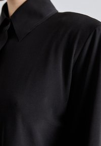 Black shirt with a structured collar, made of smooth fabric. Features loose sleeves and a subtle sheen, creating a minimalist design.