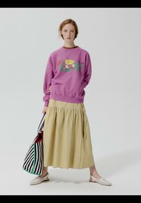 Jeune femme debout portant un sweat-shirt violet à motifs floraux, une jupe midi beige, des chaussures blanches à enfiler, tenant un grand sac fourre-tout rayé noir et blanc.