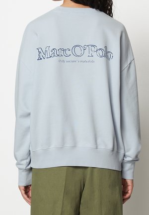 Person trägt hellblauen Pullover mit "Marc O'Polo" und "Only nature's materials" auf dem Rücken, kombiniert mit olivgrünen Hosen.