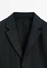 Schwarzer Leinenblazer mit Reverskragen, strukturierten Schultern und offenem Vorderteil. Die Textur ist glatt und leicht, ideal zum Schichten.