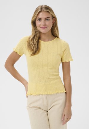 Jeune femme aux longs cheveux blonds portant un haut jaune texturé à manches courtes ajusté et un pantalon beige, souriante avec une main sur la hanche.