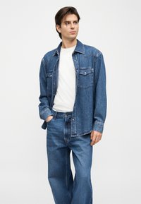 Νεαρός άνδρας που στέκεται με τα χέρια στις τσέπες, φοράει μπλε πουκάμισο από τζιν, λευκό μπλουζάκι και μπλε τζιν παντελόνι, κοιτάζοντας προς τα πλάγια σε απλό φόντο.