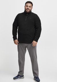 Svart fleece-genser med høy krage og halv glidelås, strukturert overflate. Kombinert med lysegrå bukser og marineblå treningssko.