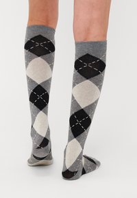 Knee-high kojinės, pagamintos iš minkšto trikotažo, su pilku, juodu ir kreminiu argyle raštu ir dekoratyvinėmis siuvimo detalėmis.