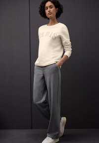 Cremefarbener Sweatshirt mit dem Schriftzug "PARIS", kombiniert mit hellgrauen, dünn gestreiften Hosen. Das Modell trägt weiße Sneakers und zeigt lockiges Haar.