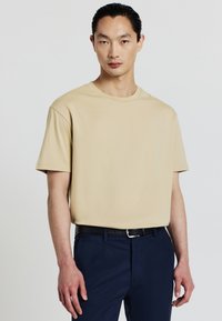 Uomo con i capelli pettinati all'indietro, che indossa una camicia beige a maniche corte infilata nei pantaloni blu navy con una cintura nera, in piedi davanti a uno sfondo semplice.