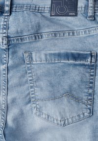 Tissu en jean bleu clair avec une poche arrière présentant un léger contour de design. Comprend des coutures visibles et une étiquette avec le nom de la marque.