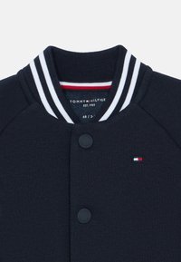 Námořnická mikina s žebrovaným límcem, který má bílé pruhy. Přední knoflíky a malá nášivka s logem v červené, bílé a modré barvě na hrudi.