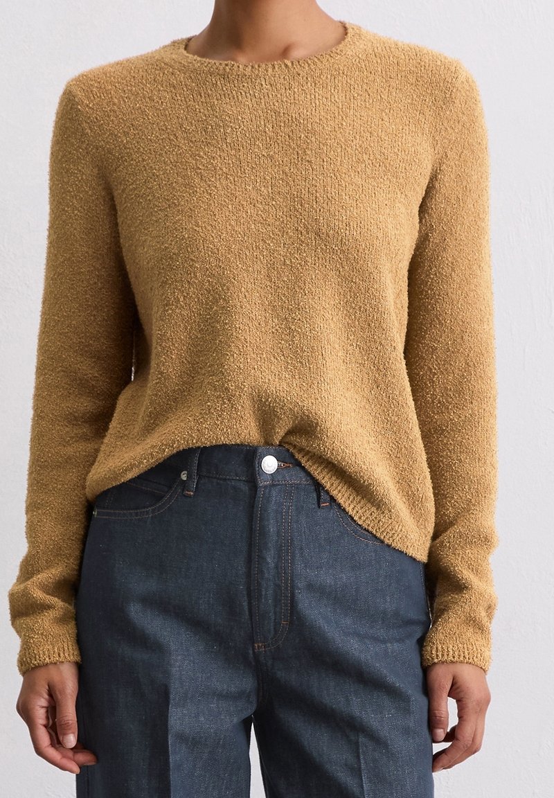 Mustard-colored knit sweater with a textured surface, round neckline, and long sleeves, paired with dark denim trousers.